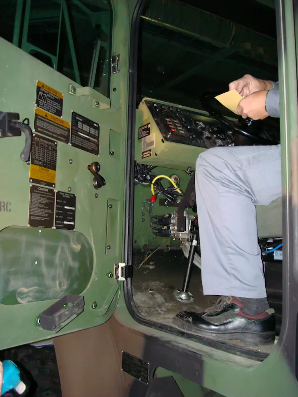 A person sitting in the driver's seat of a military truck holds a small card near the steering wheel