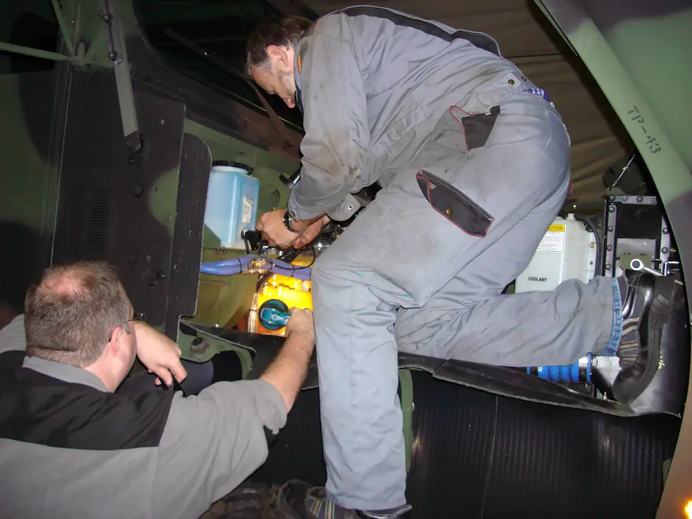 Two mechanics in gray coveralls work on the engine compartment of a vehicle