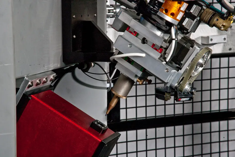 Close-up of a robotic manufacturing tool with a precision nozzle positioned above a red housing