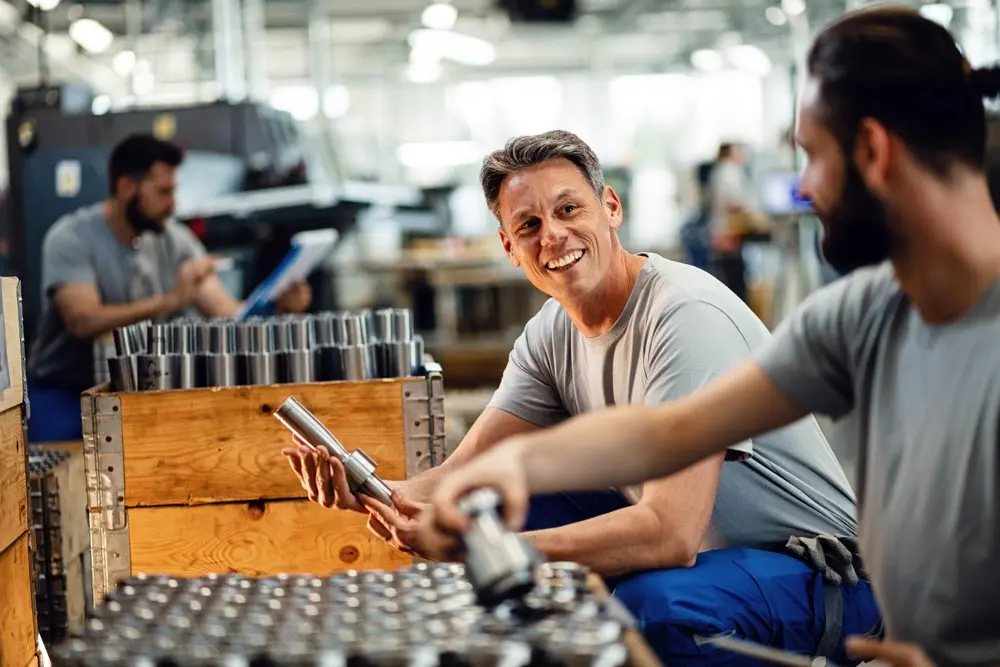 Two smiling male workers inspect metal parts in a busy factory workshop