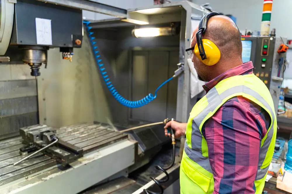 A worker wearing ear protection and a high-visibility vest operates a CNC machine in a factory