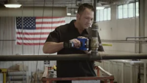 A technician uses a power tool to work on a pipe inside a building with an American flag on the wall
