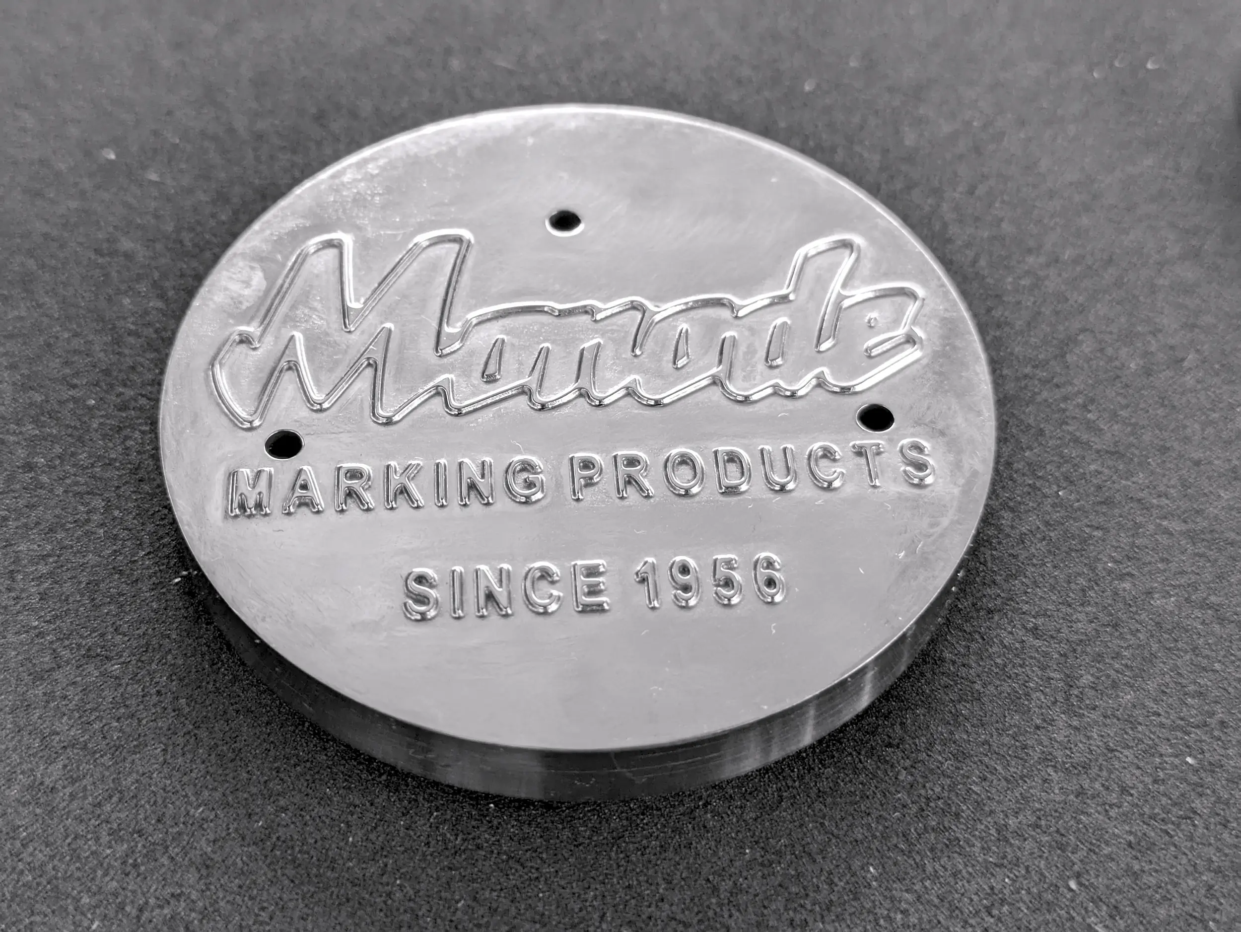 Round metal plaque embossed with the text Manade MARKING PRODUCTS SINCE 1956