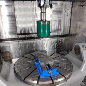 CNC milling machine interior with a green tool holder above a blue vise on a rotary table