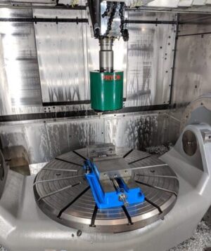 CNC milling machine interior with a green tool holder above a blue vise on a rotary table