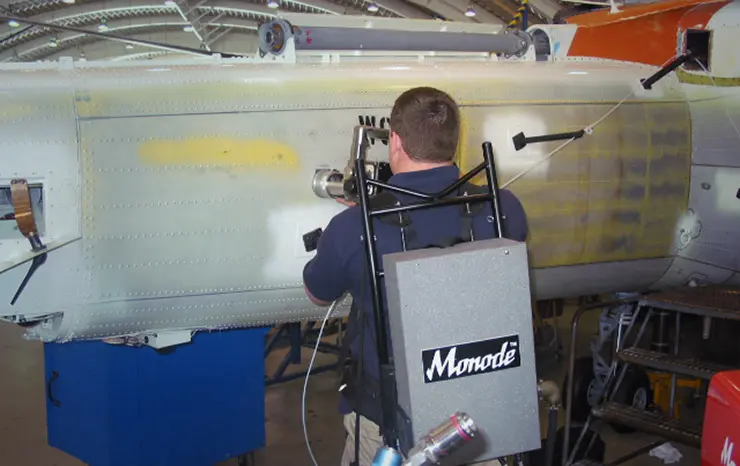 A technician wearing a Monode backpack tool uses a handheld device to inspect the exterior fuselage of an aircraft