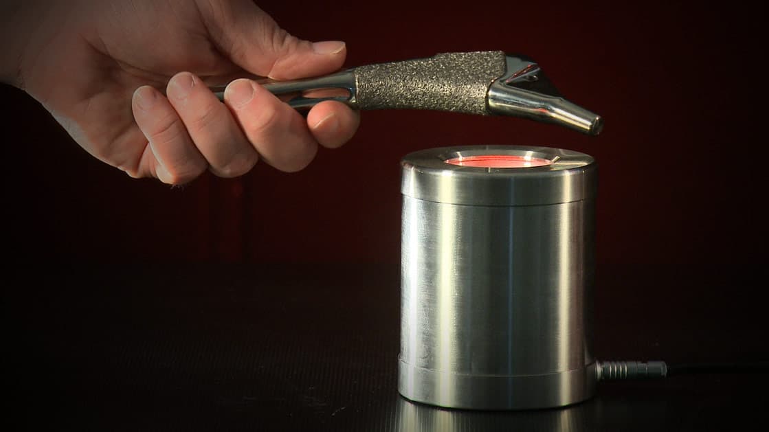 A hand holding a medical hip implant component above a heated metal cylinder