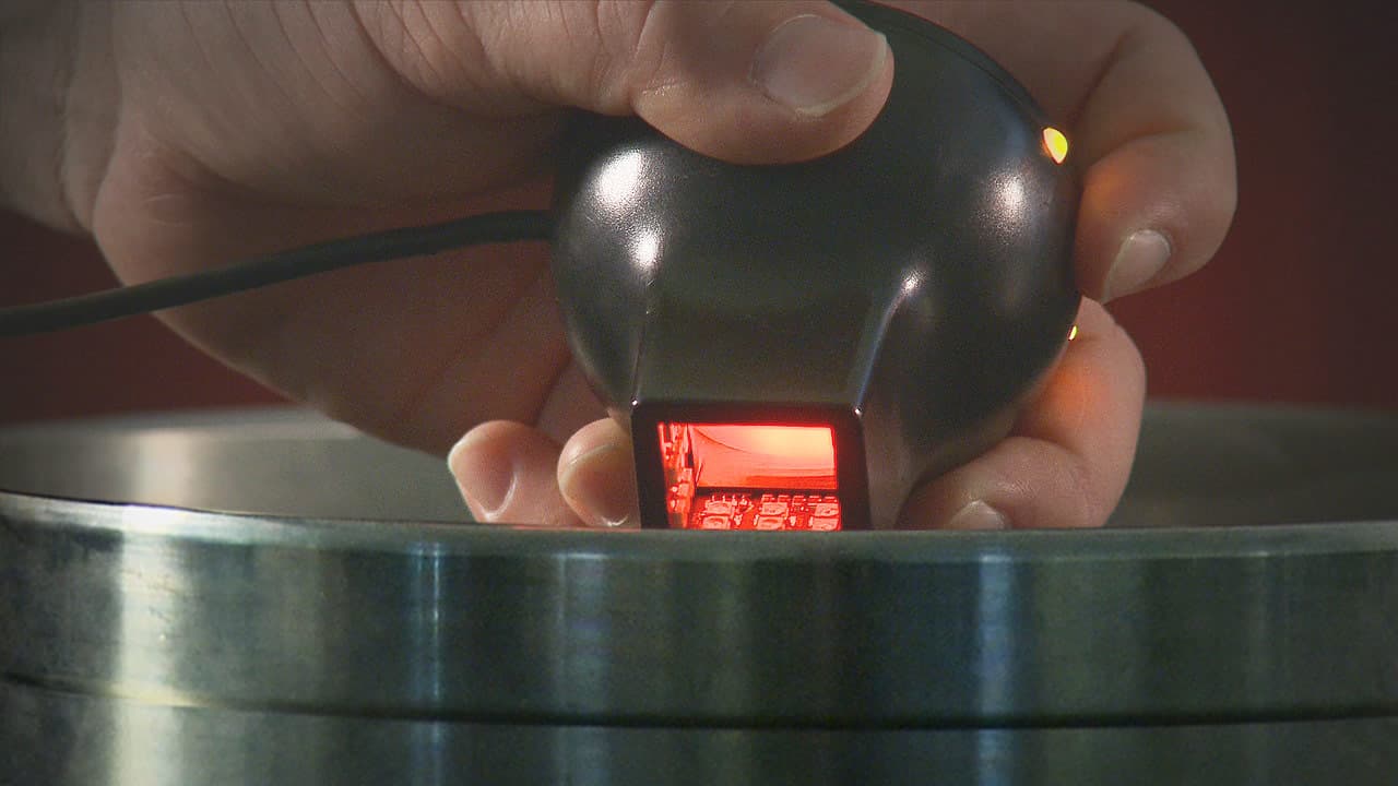 A hand holding a handheld optical scanner emitting a red light over a metal surface
